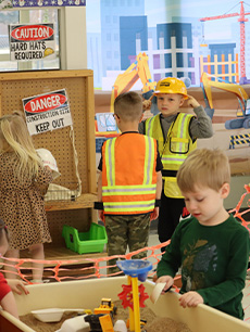 A group of children converse at a pretend construction site in the background, while in the foreground a child plays with a sandbox table