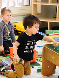 A child plays with a toy dinosaur, while another child observes