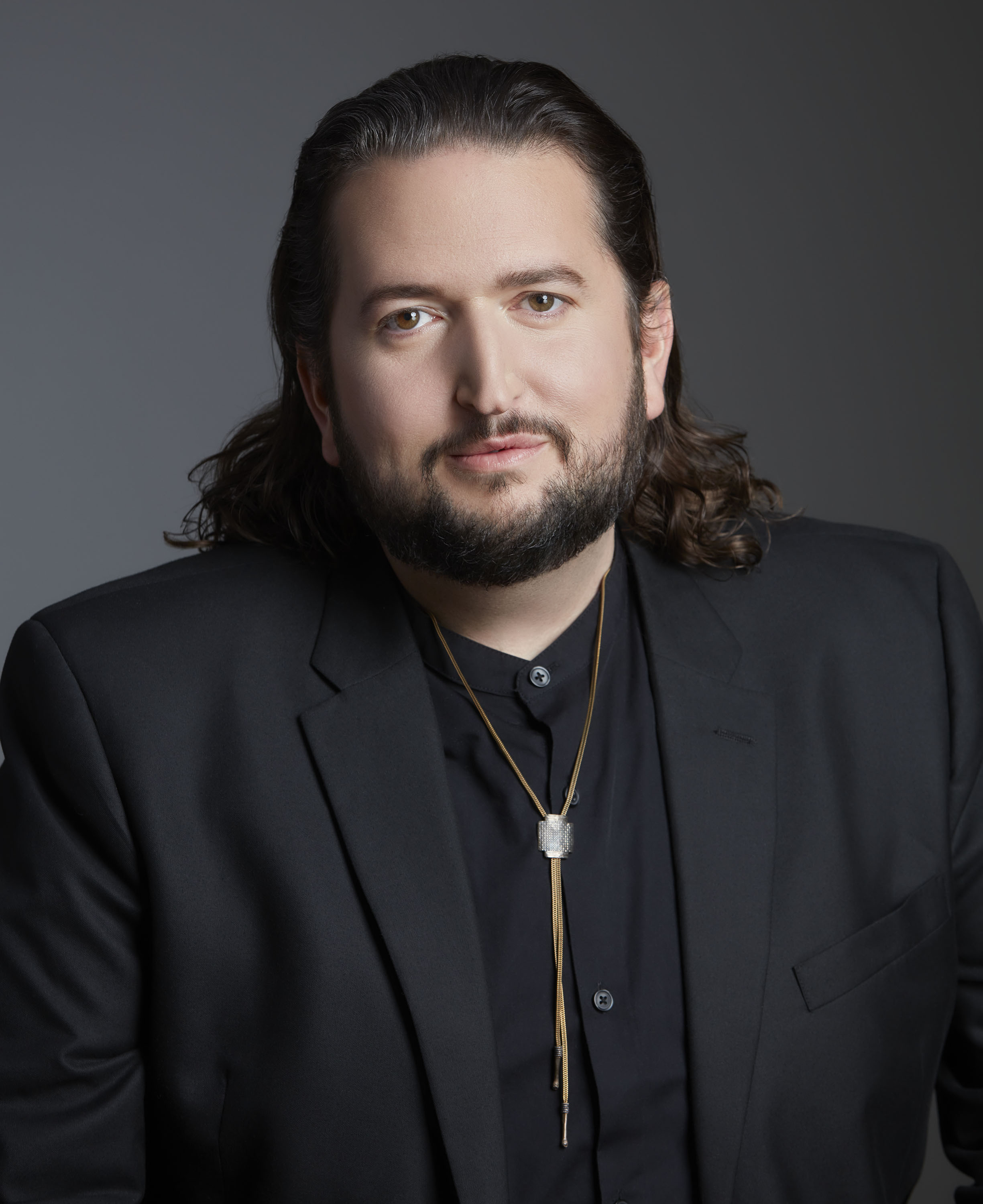 Chris Willard, a man with long dark hair and beard in a black suit in front of a grey backdrop