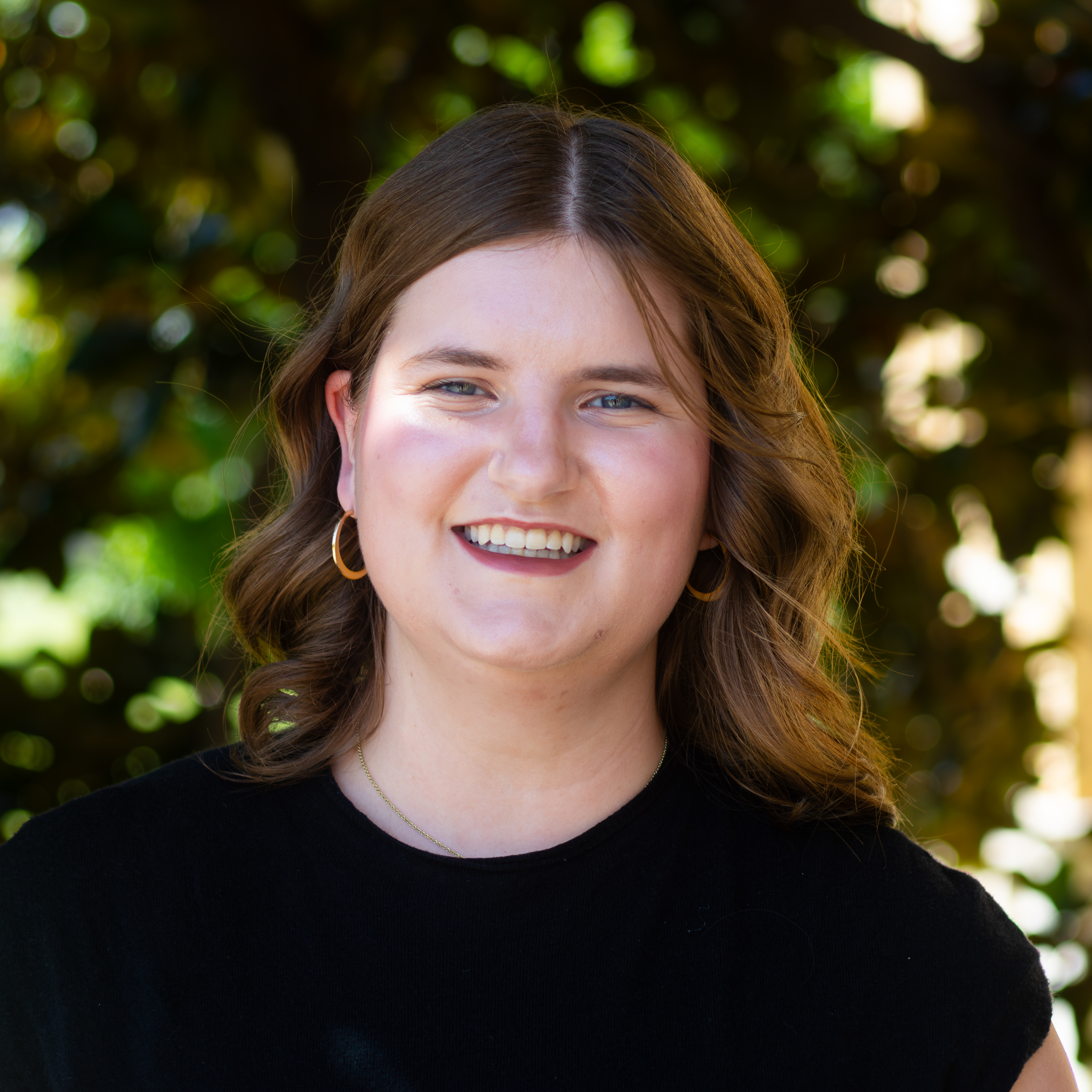 woman smiling in black t-shirt with gold earrings and wavy brown hair