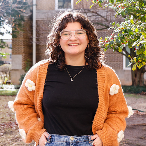 Smiling woman with octagonal glasses and wavy brown hair, in a black shirt and orange sweater, in front of green trees and a brick building