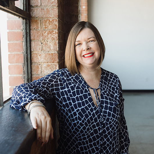 Smiling woman with brown hair, wearing a black and white patterned shirt, leans on a window ledge in front of a brick wall