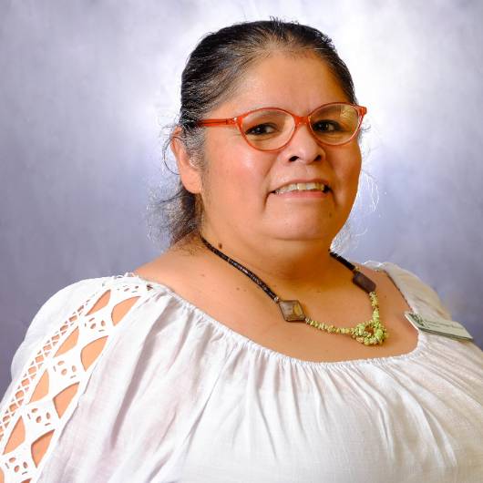 Woman wearing orange glasses and a black, tan and green necklace, with a white shirt.